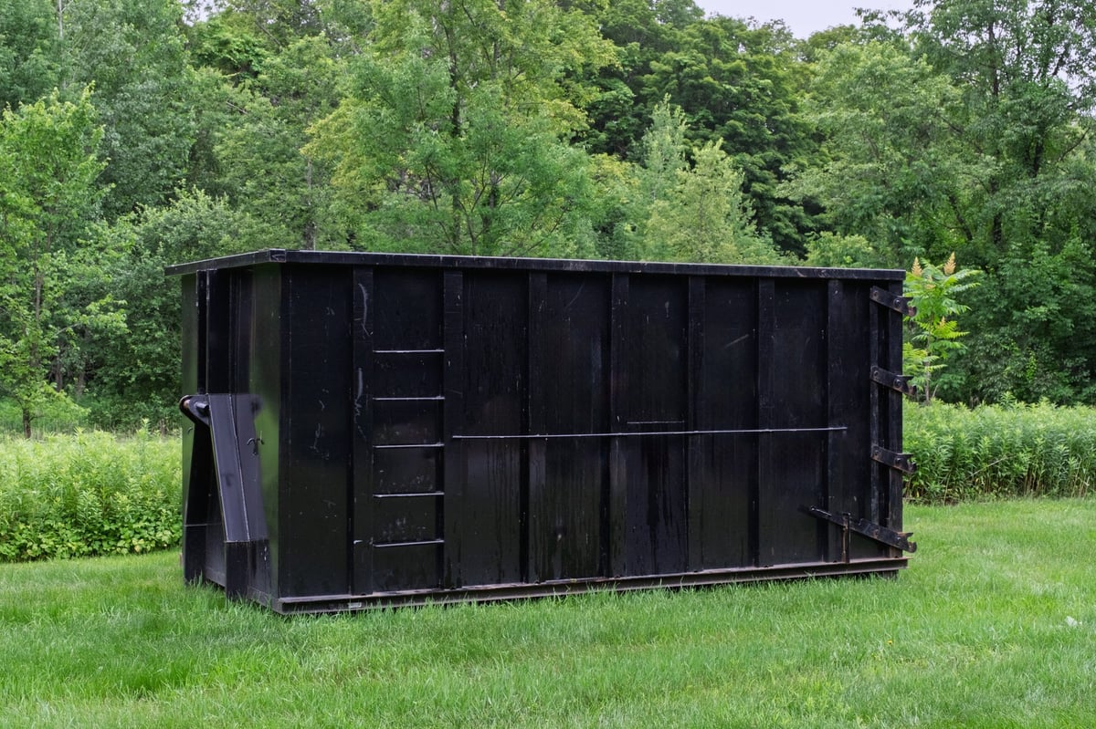 A black roll-off dumpster on a grassy lawn with trees